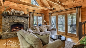A cozy cabin interior with a stone fireplace, wooden beams, a sofa, and large windows. - Sweet Water Retreat | No Pet Fees | Sleeps 5 | Coosawattee Riverfront (Ellijay)