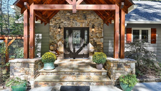 A stone porch with a wooden roof, a stone wall, and a door with glass panels. There are two potted plants on either side of the door. - Lake Reflections | Sleeps 6 | No Pet Fee | lakefront w/dock | paved | Hot Tub (Hiawassee)