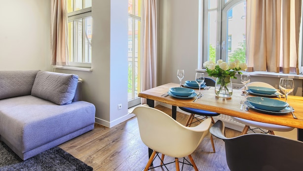 Una sala de estar moderna con un sofá, una mesa de comedor con platos azules y flores blancas, y ventanas amplias con cortinas.
