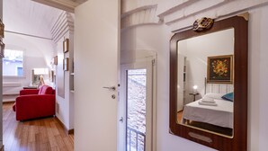 A hotel room with a wooden floor, a red sofa, a bed with a white duvet, a large mirror, and a window with a view of stone walls.