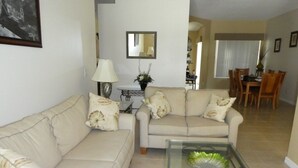 A living room with a beige sofa, a glass coffee table, and a dining area in the background. - Hampton Lakes Villa 5 Bedroom Home (Davenport)