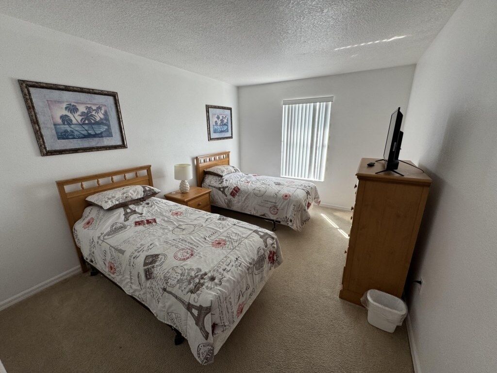 A bedroom with two beds, a wooden nightstand, a TV, and a window with blinds.