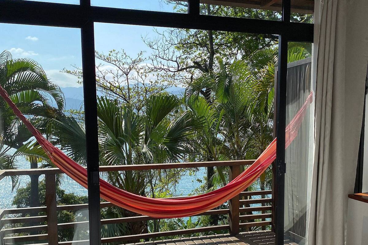 A room with a bed, a hammock, and a view of the sea and greenery.
