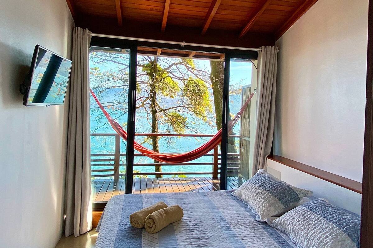 A bedroom with a bed, a TV, a hammock, and a view of the sea.