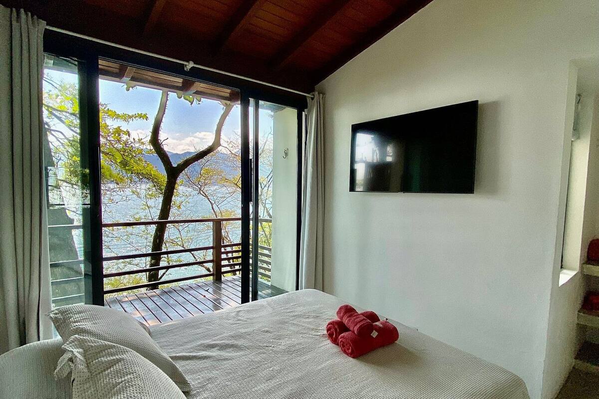 A bedroom with a bed, a ceiling fan, a TV, and a view of trees and water outside.