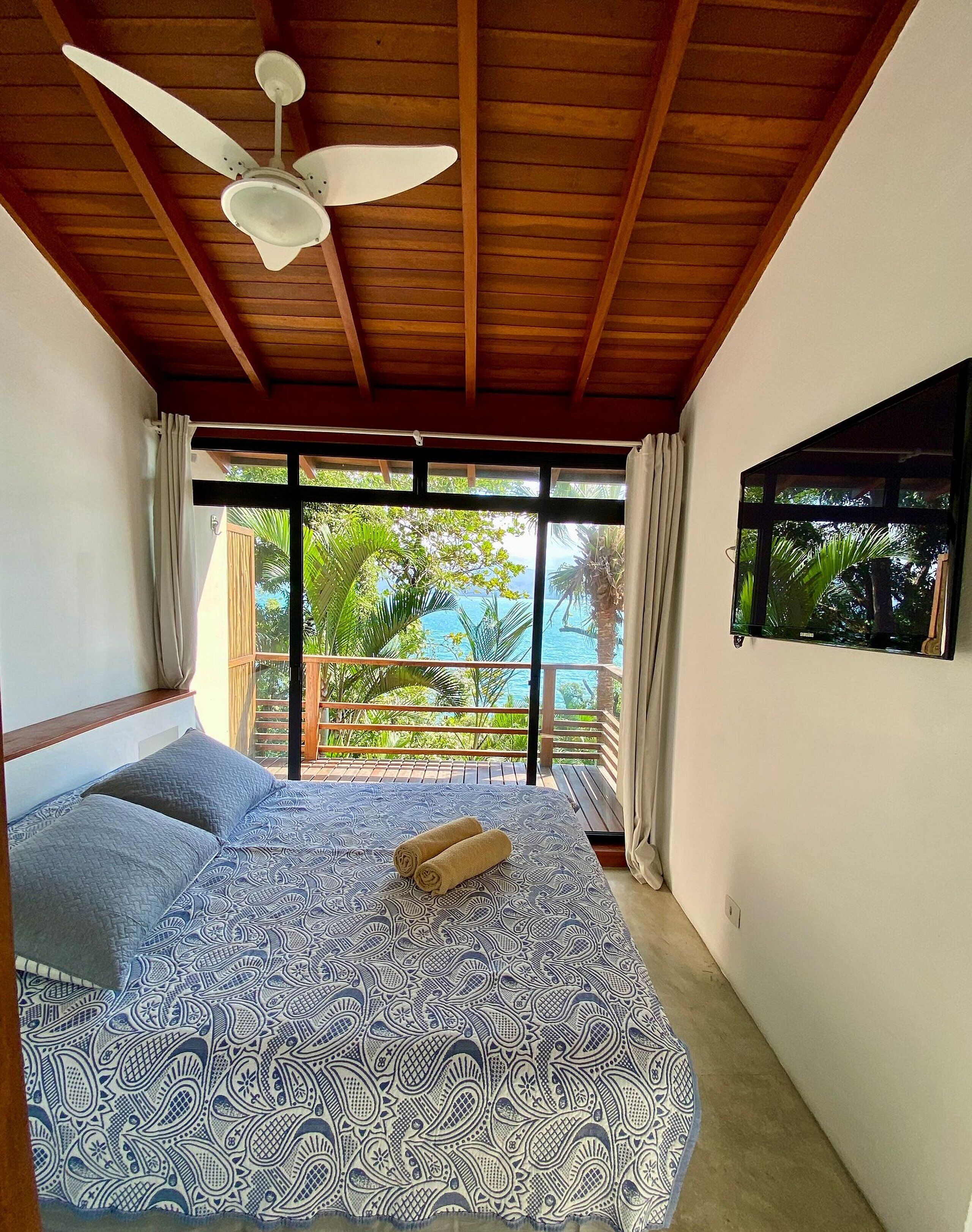 A bedroom with a bed, a ceiling fan, a window with curtains, and a view of the ocean.