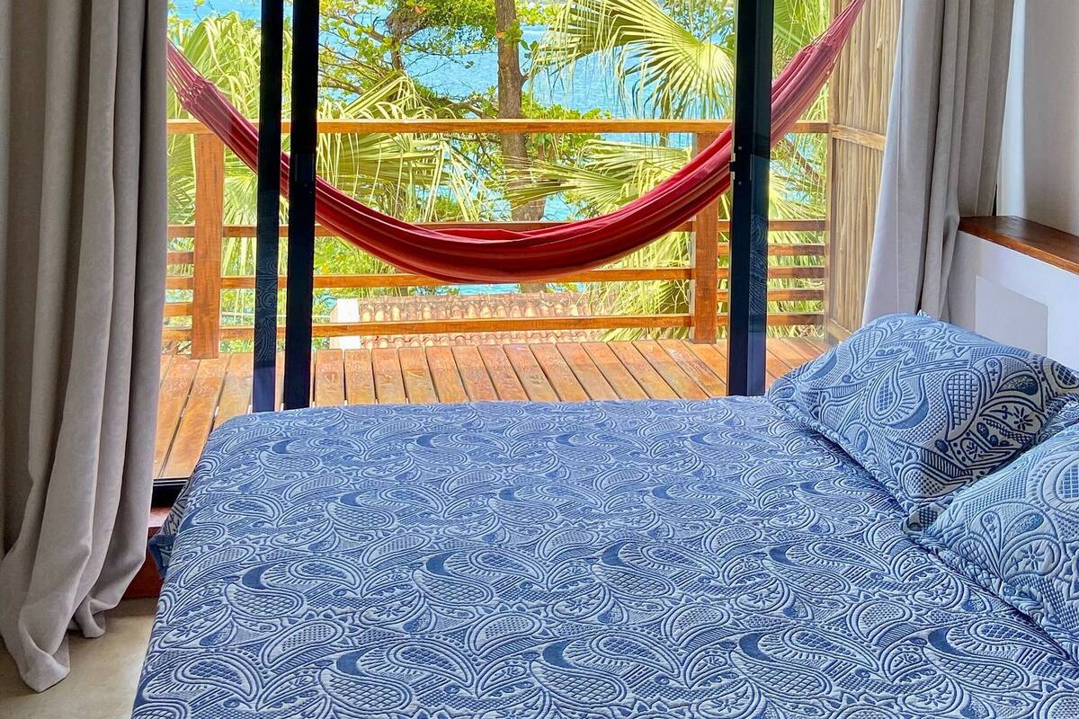 A bedroom with a large bed, a hammock, and a view of a tropical landscape through a sliding door.