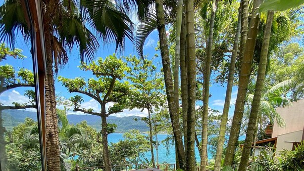 A wooden deck with a railing, surrounded by lush greenery and overlooking a body of water with mountains in the distance.