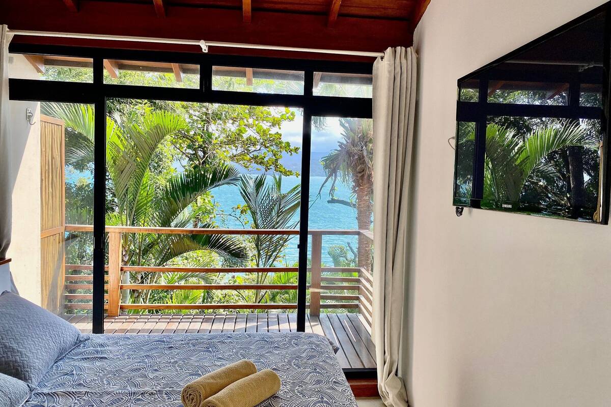 A bedroom with a large window overlooking a balcony with a view of the sea and palm trees.