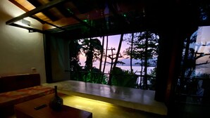 A dimly lit room with a bench, a small table, and a view of trees and a sunset through a window.