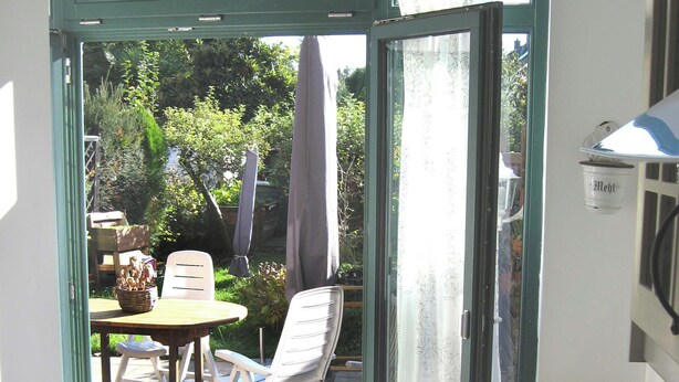 A room with a sliding glass door leading to an outdoor patio area. Inside, there is a table with a chair, a wooden chair, and a small potted plant. - 15 min from Neuss main station. Apartment for rent in quiet residential street. (Neuss)