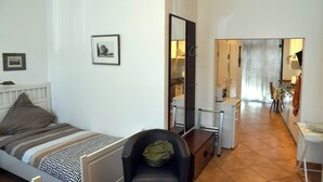 A compact bedroom with a single bed, a chair, and a mirror. A small kitchenette is visible in the background. - 15 min from Neuss main station. Apartment for rent in quiet residential street. (Neuss)