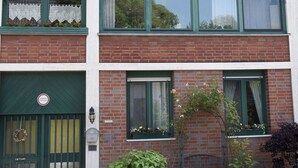 A two-story brick building with a green door and windows, a small garden with plants, and a wreath on the door. - 15 min from Neuss main station. Apartment for rent in quiet residential street. (Neuss)