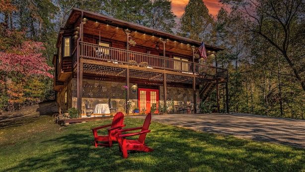 A rustic cabin with a stone facade, a balcony, and a porch with red Adirondack chairs.