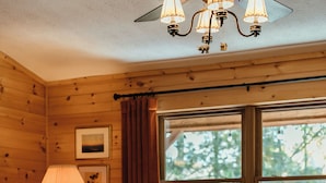 A cozy cabin interior with a wooden interior, a large window with a view of trees, a table with books, and a lamp.
