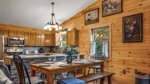 A wooden cabin interior with a kitchen, dining area, and a large window.