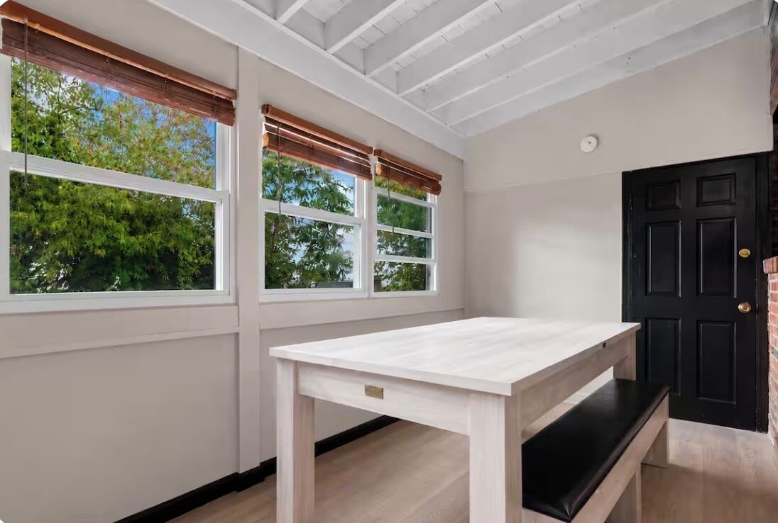 Une pièce avec une table blanche, deux fenêtres équipées de stores, une porte noire et une horloge bien visible.