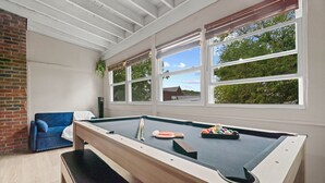 A pool table with cues and balls, a blue sofa, and large windows with a view of greenery.