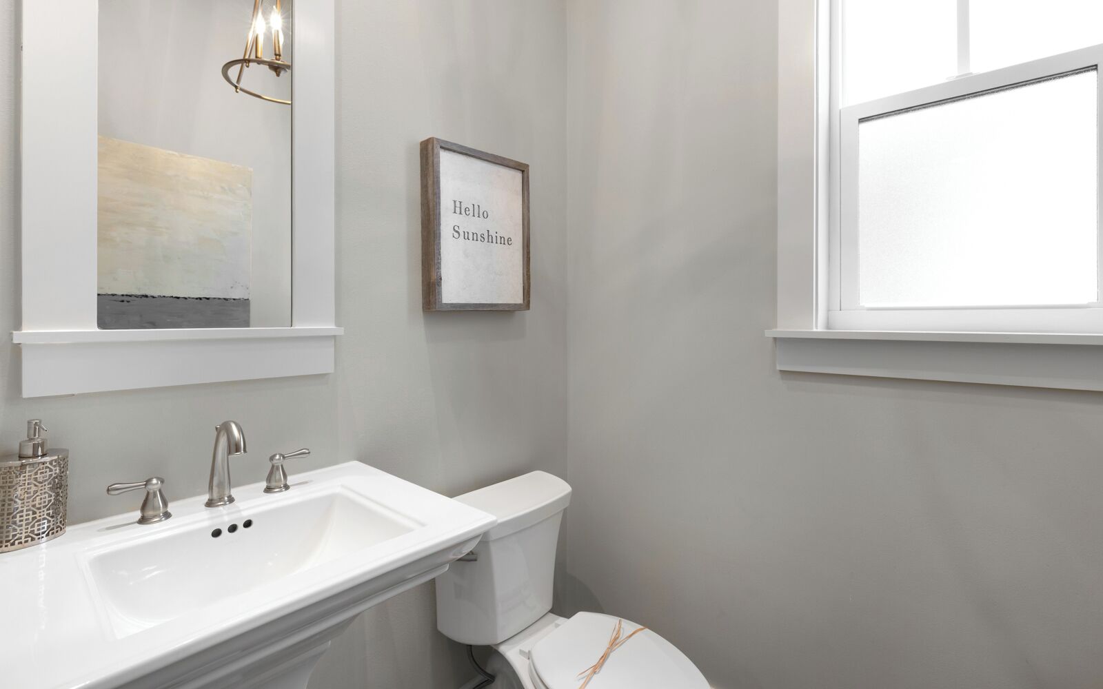A bathroom with a white sink, two faucets, a toilet, and a framed sign that reads "Hello Sunshine."