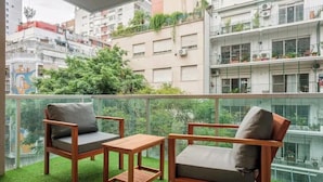 Un balcón con dos sillas de madera y una mesita pequeña, rodeado de hierba verde exuberante y vistas de edificios cercanos.