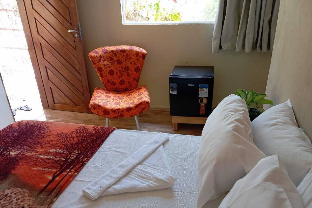 Un lit avec du linge de lit blanc, un plaid à motifs, une chaise à décor floral, une petite boîte noire et une fenêtre avec des rideaux.