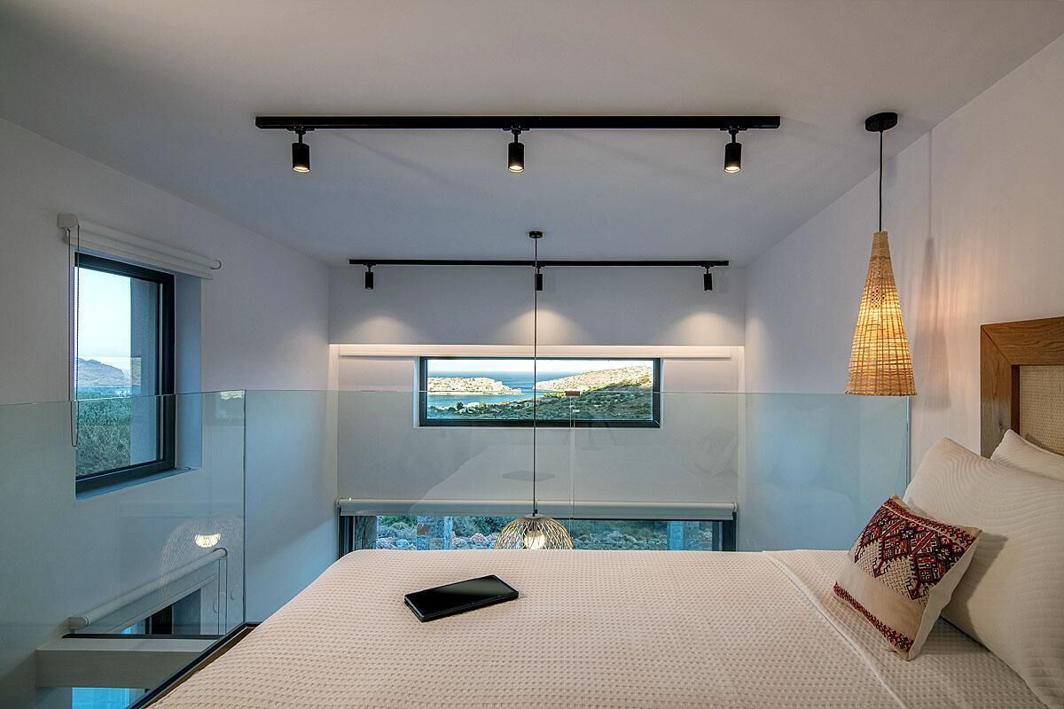 A modern bedroom with a large bed, a tablet on the bed, a view of the sea through a window, and a hanging light fixture.
