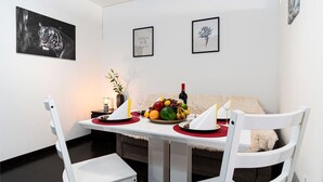 A dining area with a white table set for two, featuring a fruit bowl, wine, and glasses. There are two framed pictures on the wall, a white chair, and a sofa in the background.