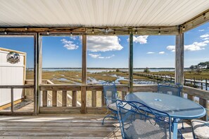 A covered outdoor area with a wooden deck, metal chairs, and a table. It features a view of a coastal landscape with a wooden fence and a distant shoreline. - Pony Swim Crossing by Seaside Vacations & Sales (Chincoteague Island)