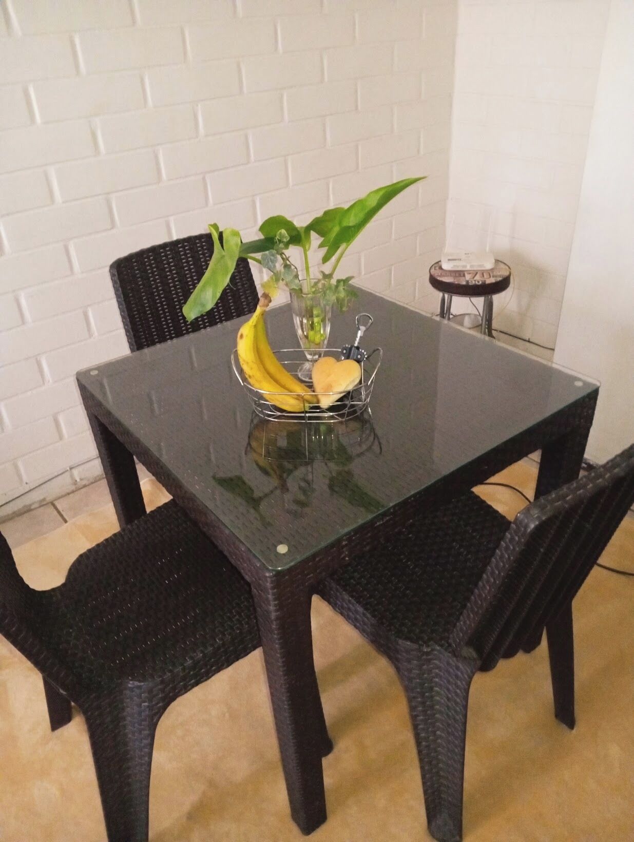 A dining table with a glass top, surrounded by black wicker chairs. A small plant with green leaves and a banana are placed on the table.