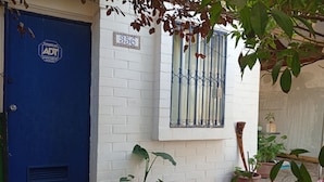 Une porte bleue avec une plaque portant un numéro, un mur en briques blanches, une fenêtre avec des barreaux en métal et des plantes en pot.