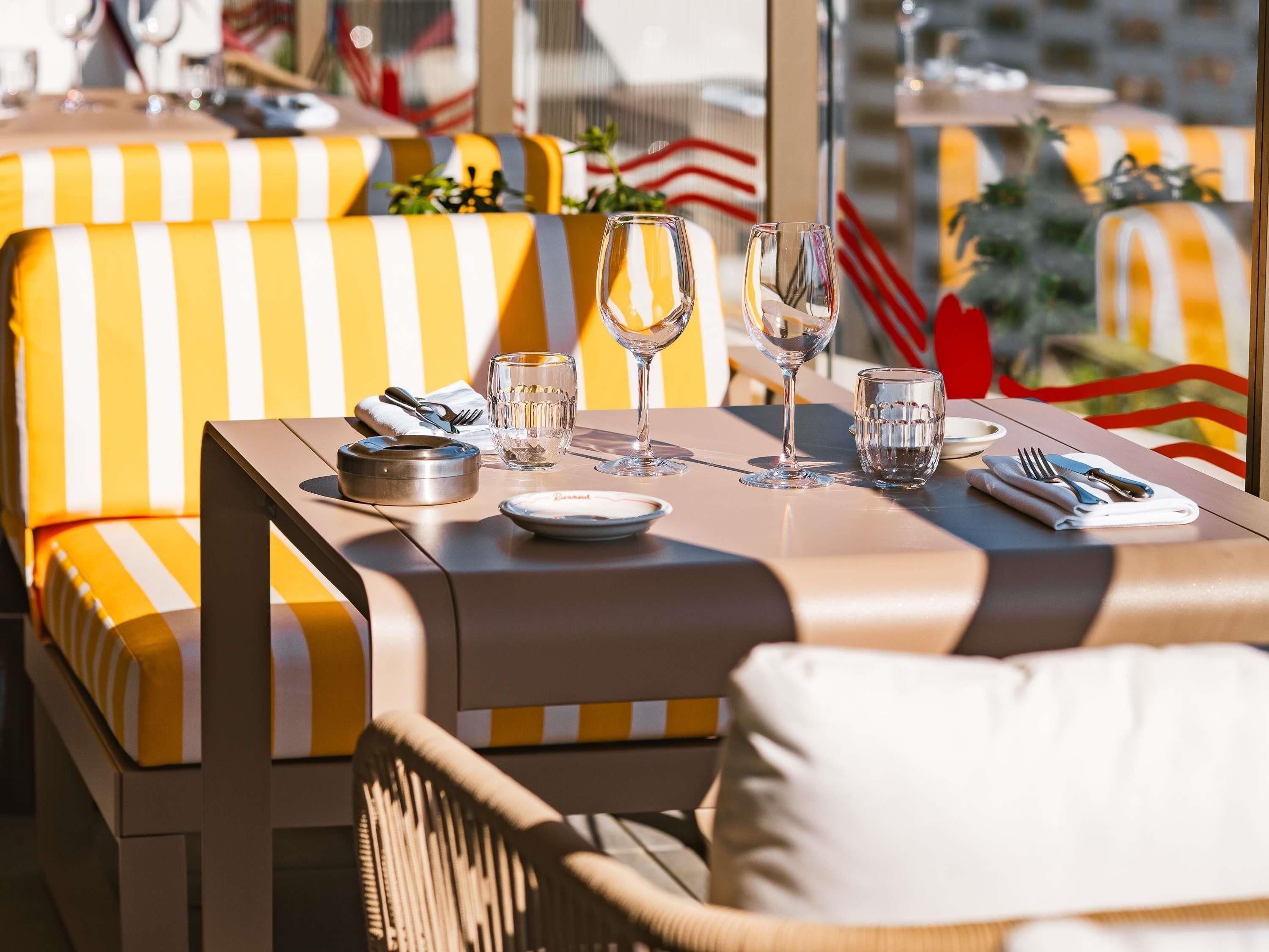 Un comedor al aire libre iluminado por el sol, con asientos a rayas amarillas y blancas, una mesa dispuesta con platos, vasos y cubiertos.