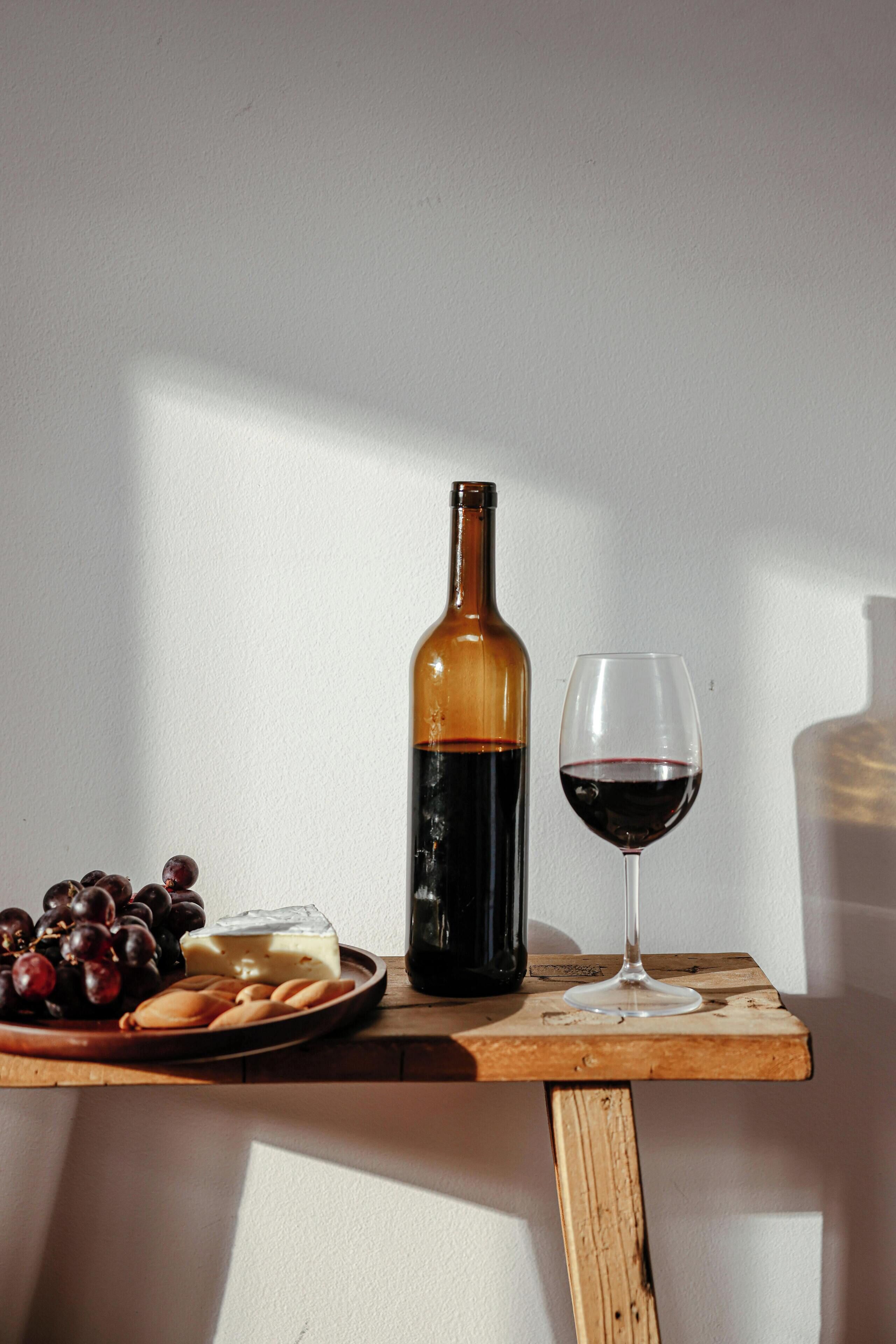 A wooden table with a bottle of wine, a glass of wine, grapes, and cheese.