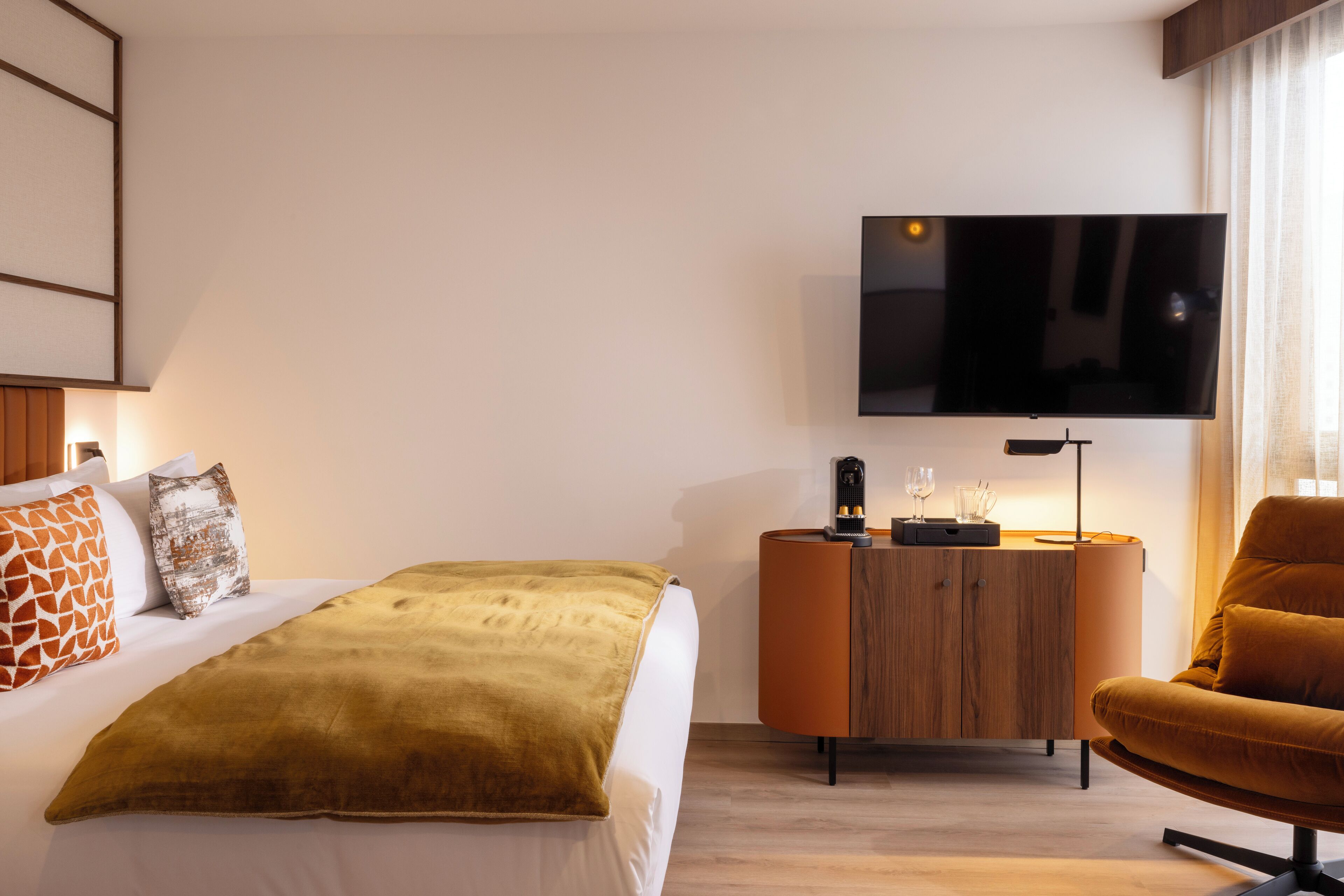 A modern hotel room with a bed, a TV mounted on the wall, a wooden side table, and a comfortable armchair.