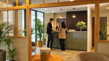 Un espace d’accueil comprenant un comptoir de réception, des plantes en pot et un coin salon avec un pouf rond.