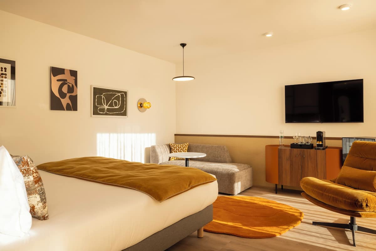 A modern hotel room with a bed, a sofa, a chair, a small table, and a TV mounted on the wall.