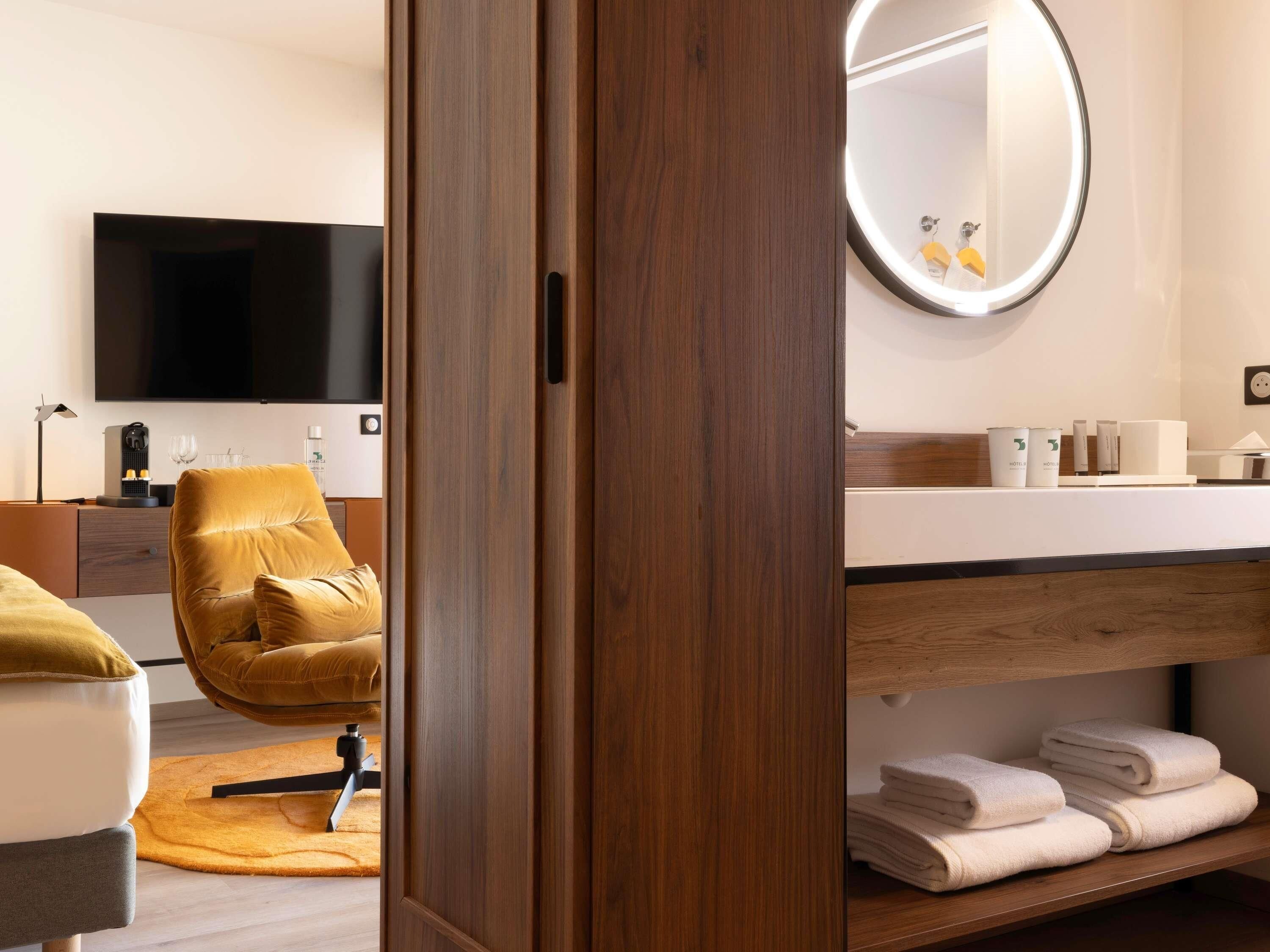Habitación de hotel moderna con puerta de madera, un espejo grande, un televisor de pantalla plana, un sofá, una cafetera y un perchero.
