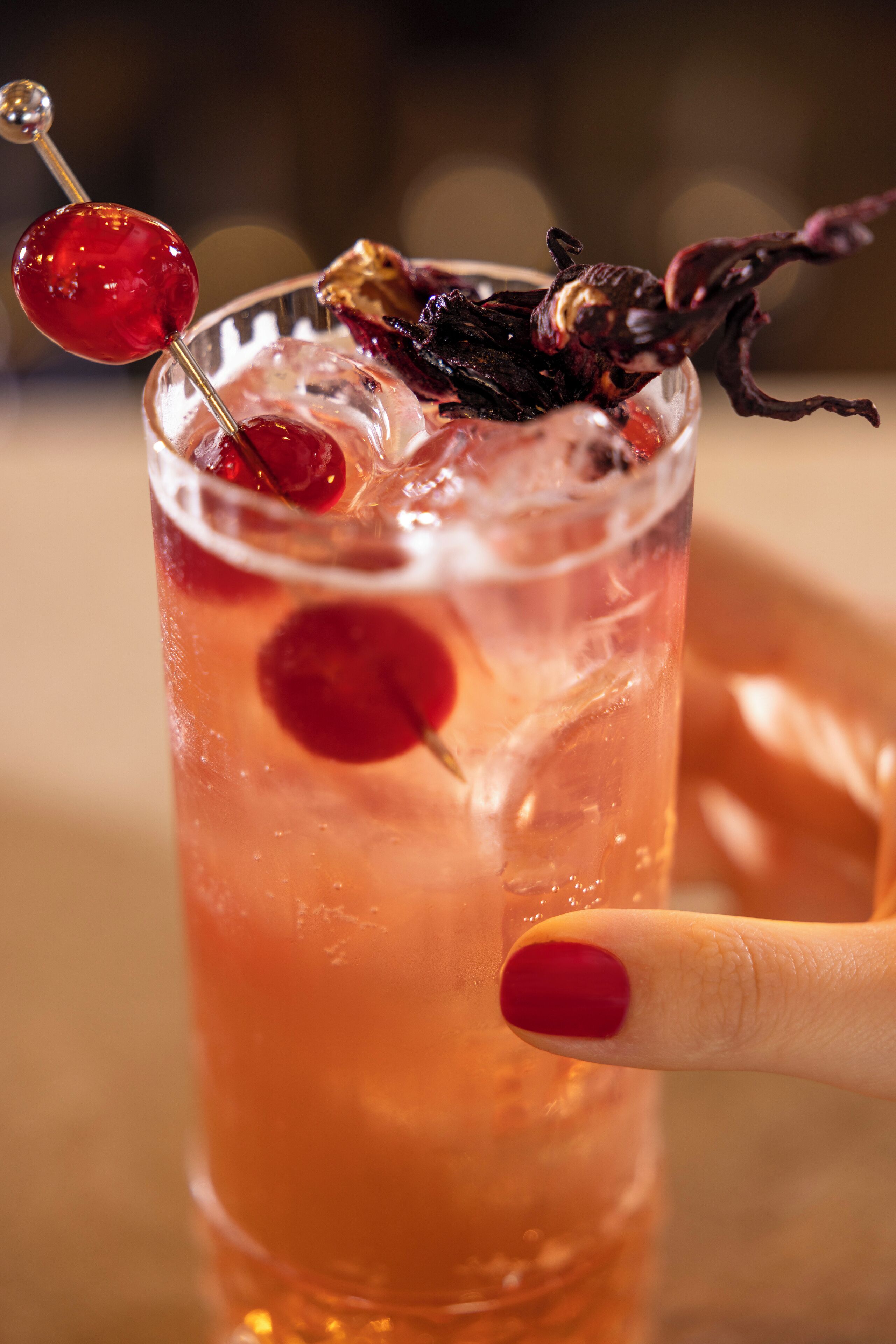 A cocktail glass with a cherry garnish and a decorative skewer, held by a hand with red nail polish.