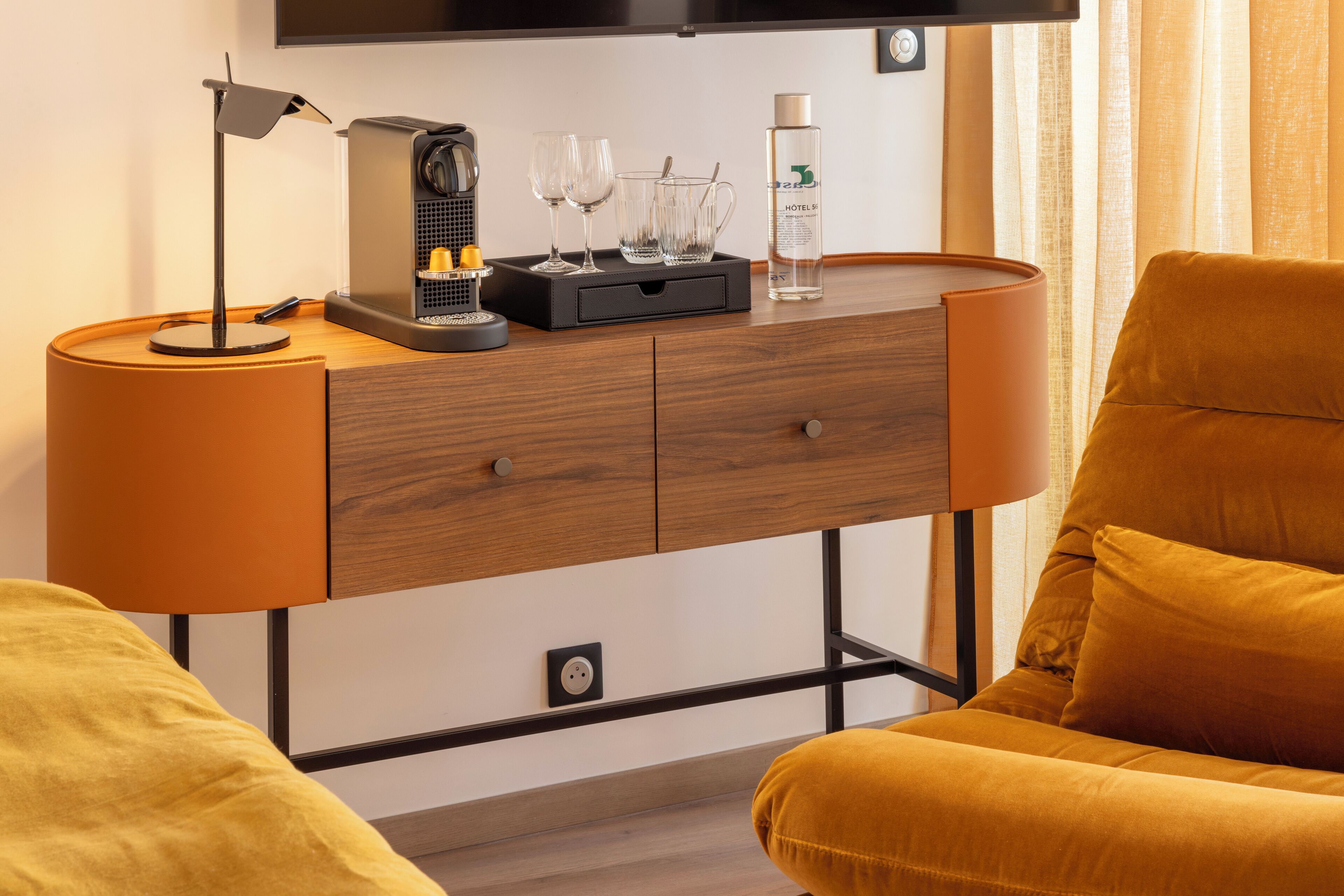 A modern hotel room with a wooden desk, a lamp, a coffee machine, and a bottle of hotel water.