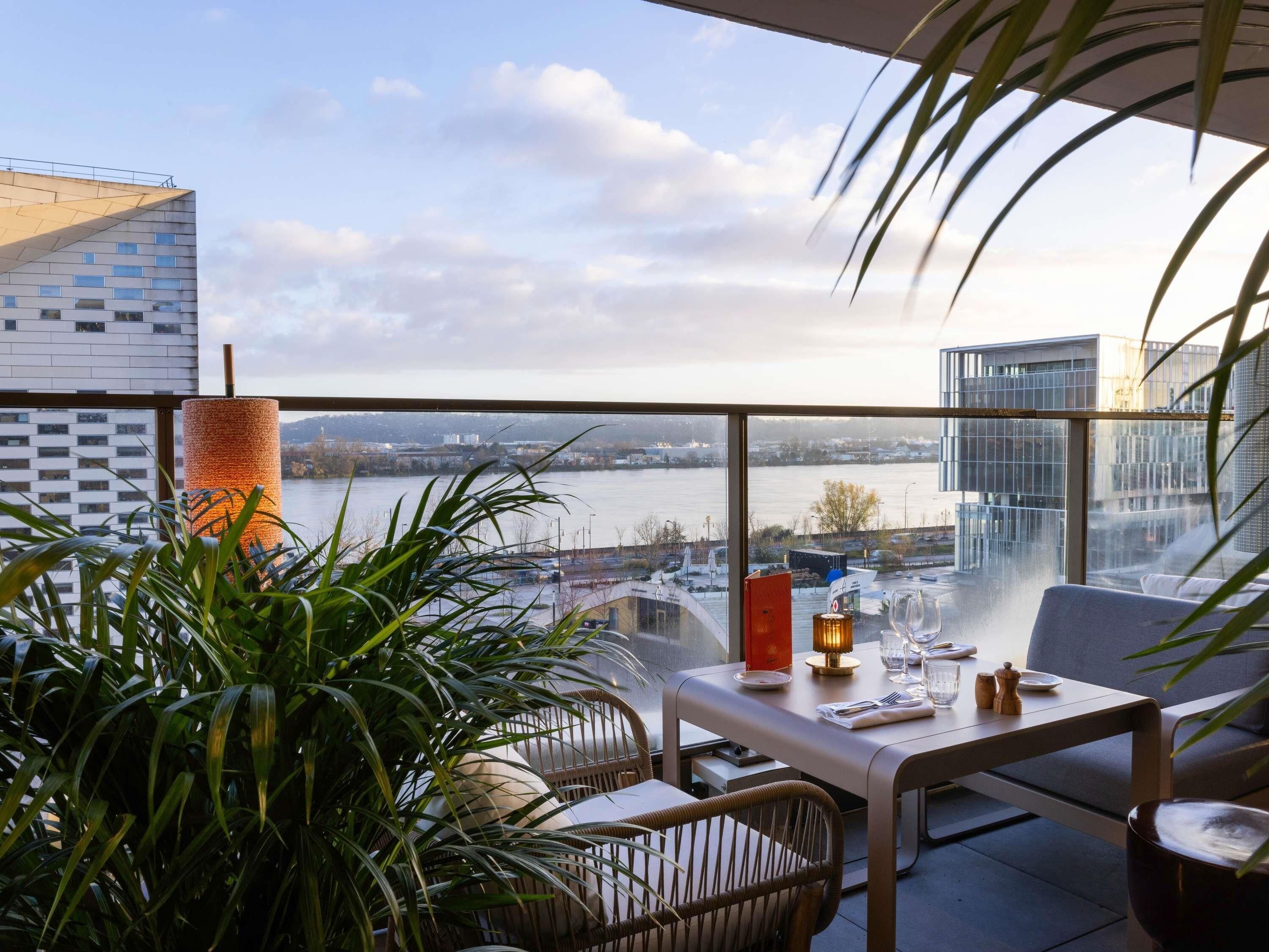 A balcony with a view of a river, a table with glasses and a candle, and potted plants.