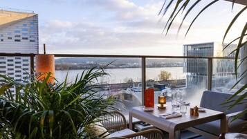 A balcony with a view of a river, a table with glasses and a candle, and potted plants.