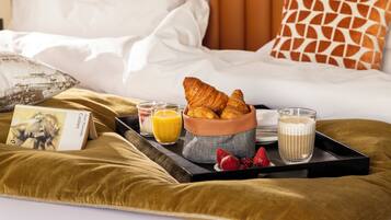 Bandeja de desayuno con cruasanes, fresas, zumo de naranja y un vaso de leche sobre una cama con un libro y una almohada.