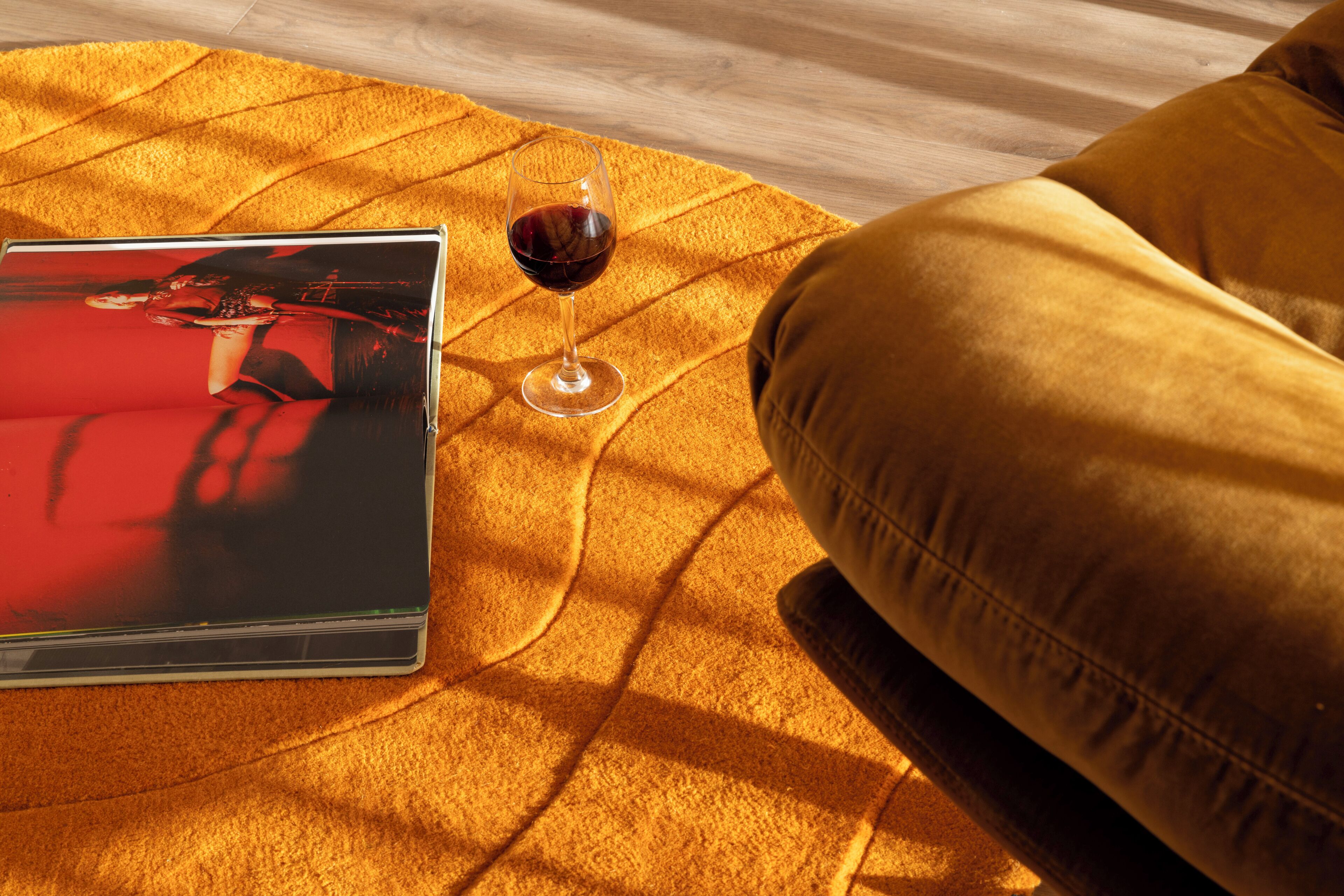 A magazine on a yellow rug, a glass of red wine, and a yellow couch.
