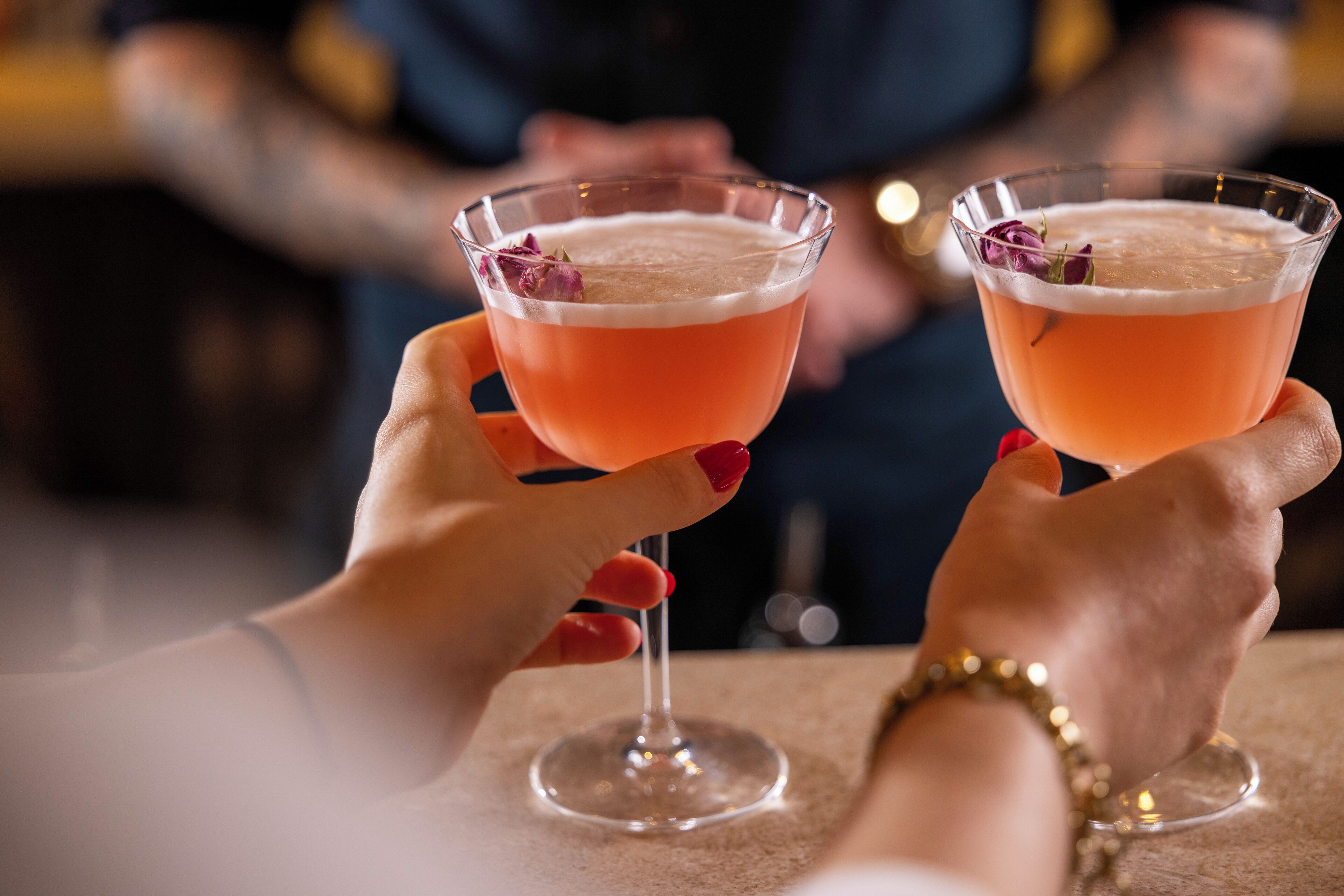 Zwei Hände halten Cocktailgläser mit einer Rosengarnitur.