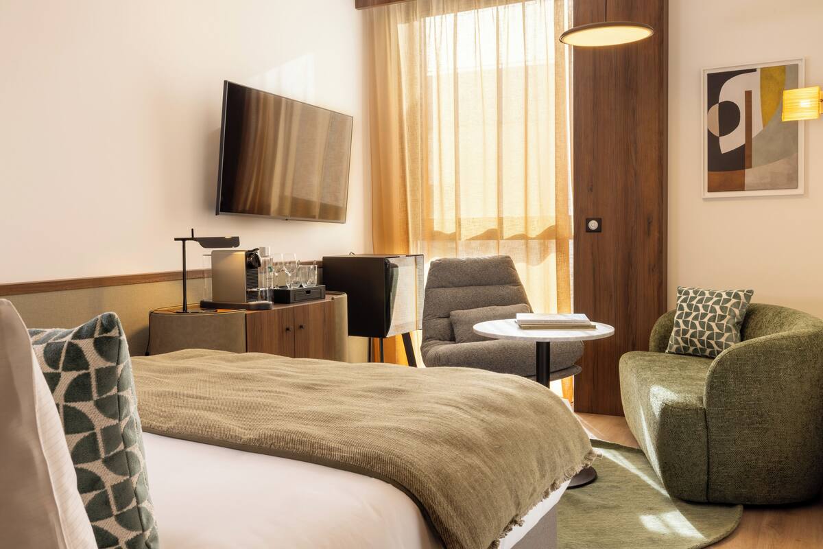 A hotel room with a bed, a sofa, a chair, a small table, a TV, and a window with curtains.
