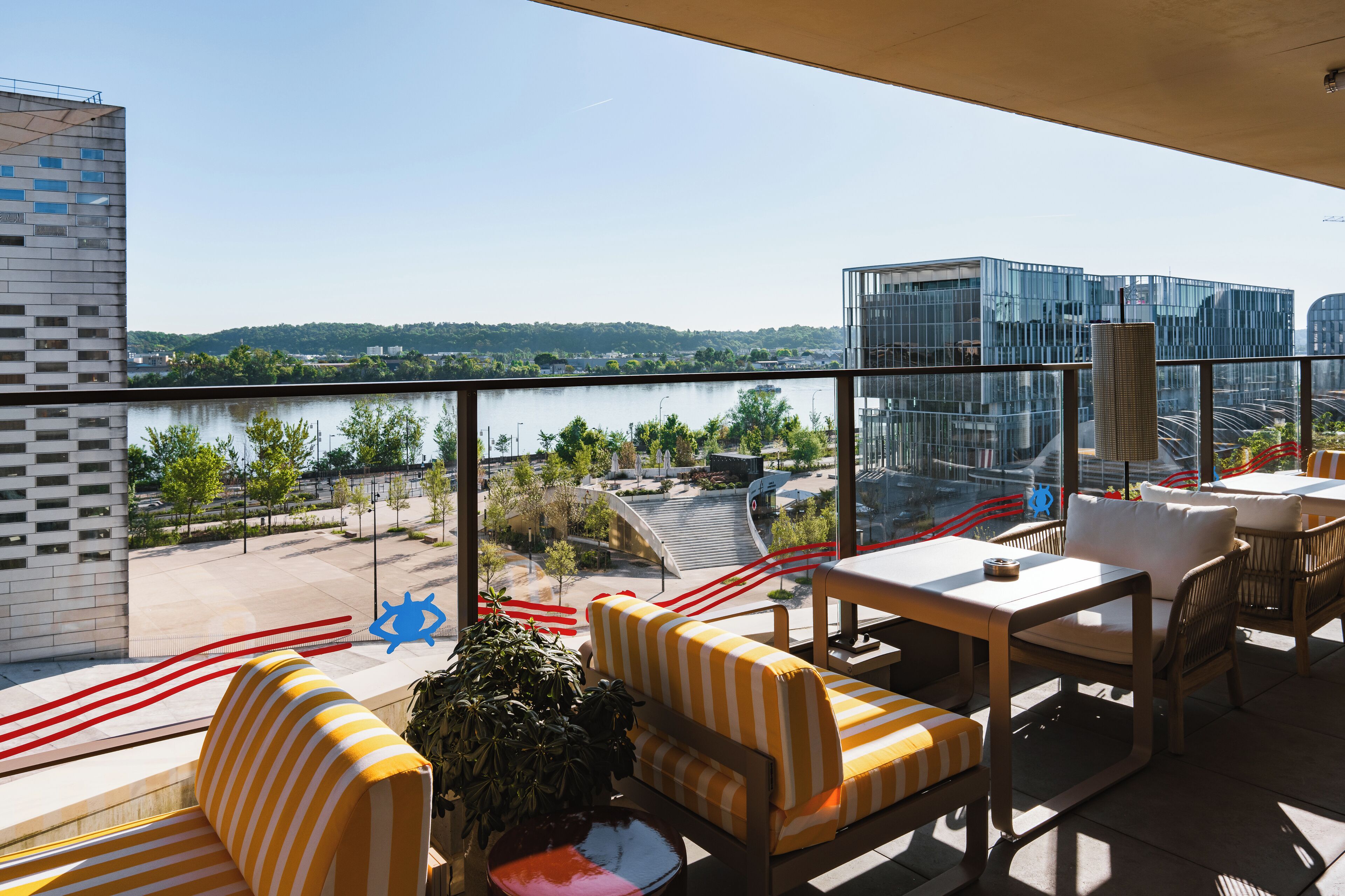 A balcony with a view of a river and modern buildings, furnished with striped sofas and a table.