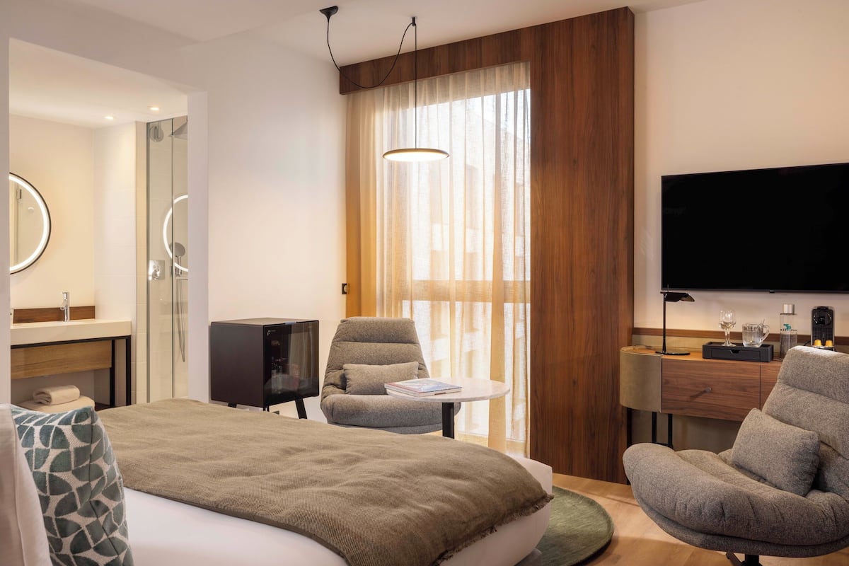 A modern hotel room with a bed, a seating area with a chair and a small table, a TV, a mirror, and a large window with curtains.