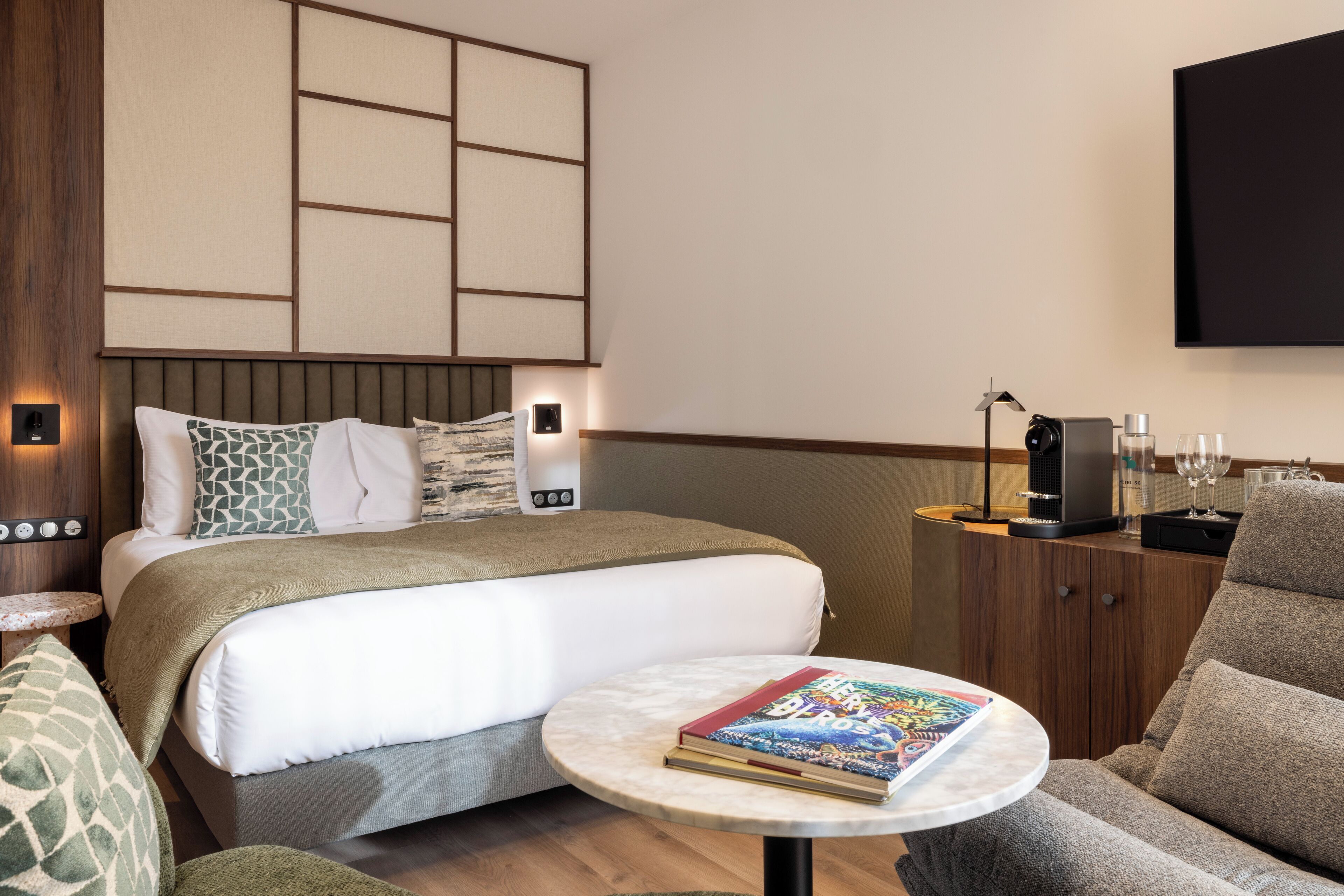 A modern hotel room with a neatly made bed, a round coffee table with a book, a sofa, a TV mounted on the wall, and a bedside table with a lamp and a coffee machine.