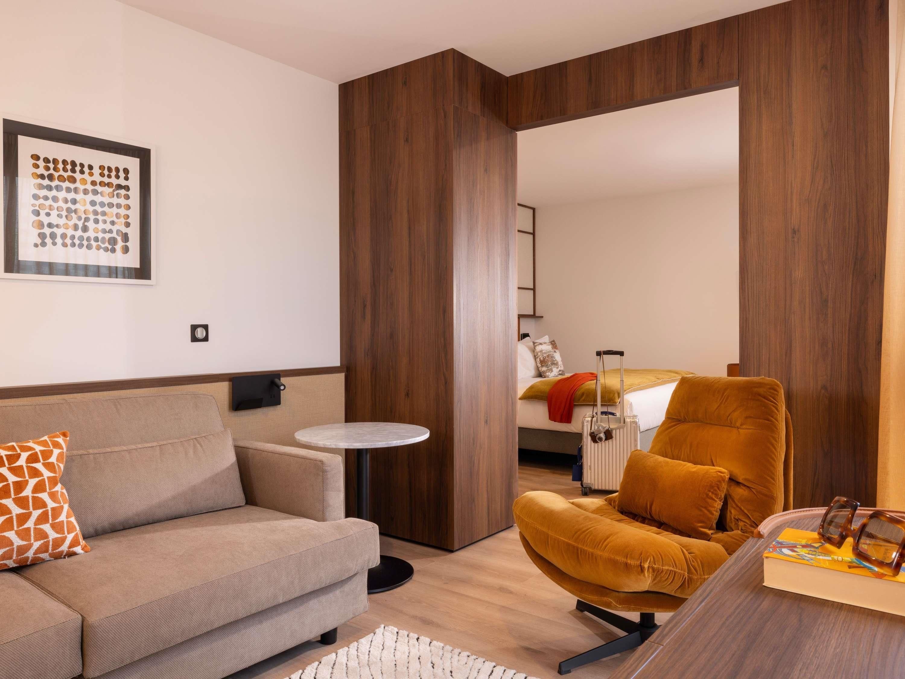 Une chambre d’hôtel moderne avec un canapé, un fauteuil, une petite table ronde et un lit sur lequel se trouve une valise.
