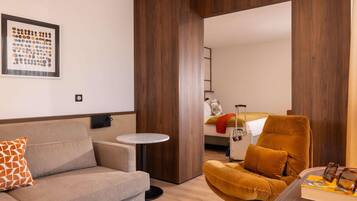Une chambre d’hôtel moderne avec un canapé, un fauteuil, une petite table ronde et un lit sur lequel se trouve une valise.