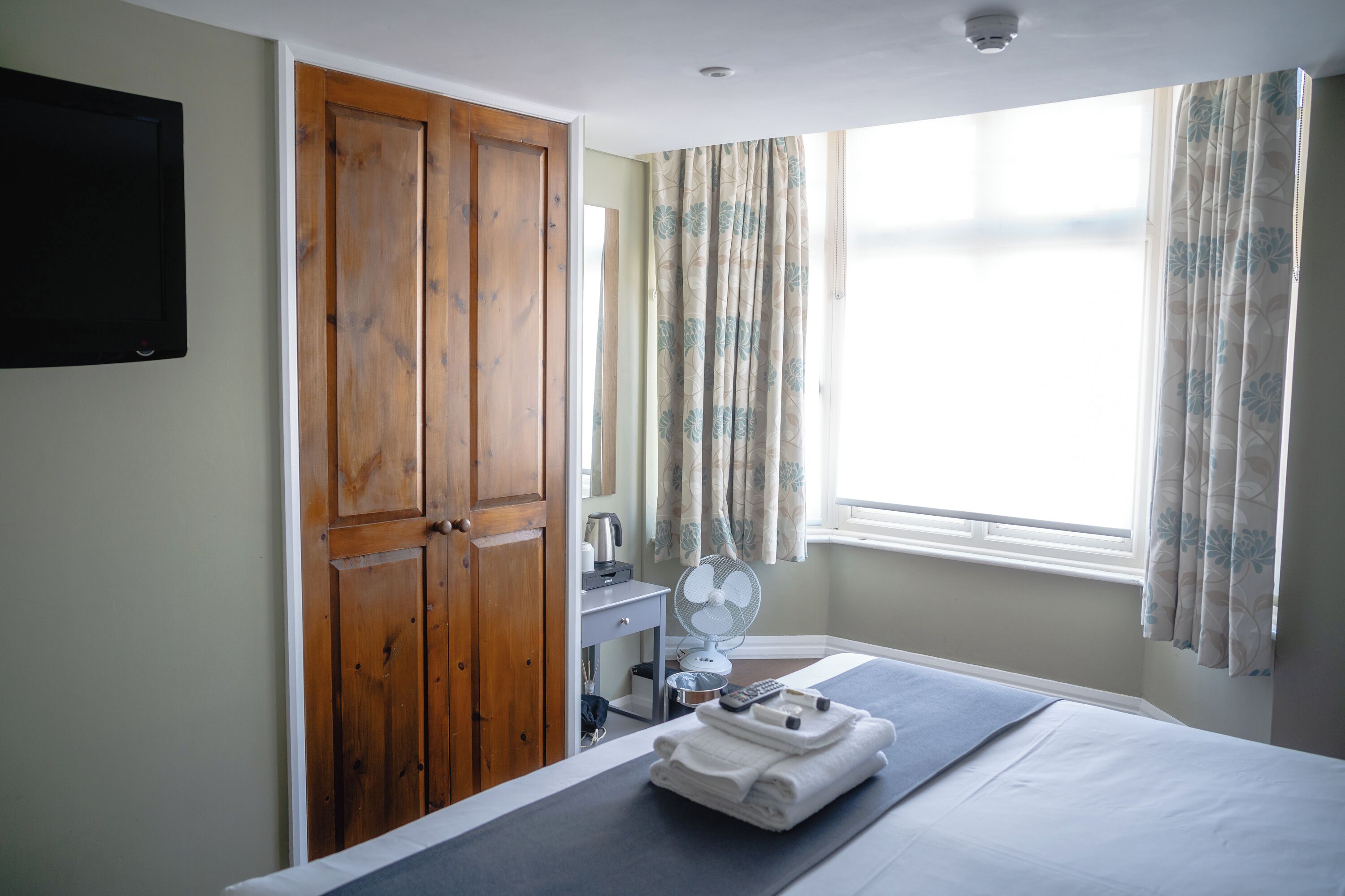 A hotel room with a bed, a wooden wardrobe, a desk with a fan, and a window with curtains.